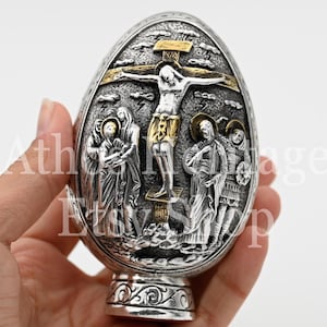 May include: Silver and gold egg-shaped ornament with a detailed depiction of the crucifixion of Jesus Christ. The ornament features a cross with Jesus on it, surrounded by three figures. The ornament has a decorative base.