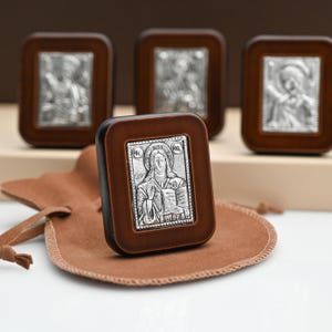 May include: Silver religious icon in a dark brown wooden frame. The icon depicts a religious figure holding a book. A brown leather pouch is in the foreground. Three additional icons are in the background.