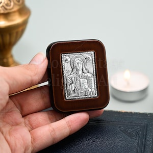 May include: A small wooden frame with a silver icon of Jesus Christ holding a book. The icon is on a dark brown background and has the letters "IC XC" and "NI KA" on the top and bottom.