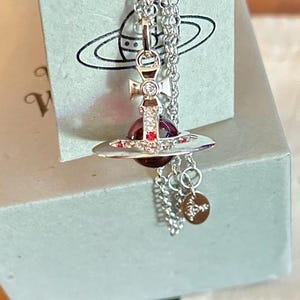 Puede incluir: Collar plateado con un colgante con un diseño de orbe rojo y cruz. El colgante cuelga de una delicada cadena, con un pequeño dije adjunto. El collar se exhibe en una caja azul claro.