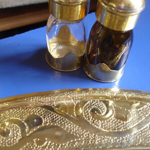 Op de afbeelding: Twee glazen strooiers met gepolijste goudkleurige metalen doppen en voetjes, geplaatst achter een grote, ronde schaal van gehamerd messing met een ingewikkeld, reliëf en getextureerd golfachtig patroon langs de brede, gebogen en reflecterende rand.