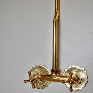 Può includere: Un rubinetto dorato con due maniglie ornate. Le maniglie sono rotonde e hanno un design floreale decorativo. Il rubinetto è montato su una parete bianca.