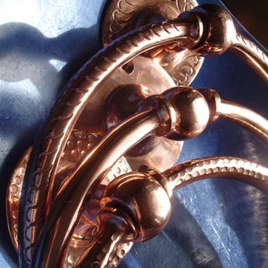Könnte beinhalten: Nahaufnahme eines Satzes von Türgriffen aus Kupfer. Die Griffe haben ein geschwungenes Design mit dekorativen Details und einer polierten Oberfläche. Die Hardware ist auf einer blauen Oberfläche montiert, wobei sich das Licht im Kupfer spiegelt.