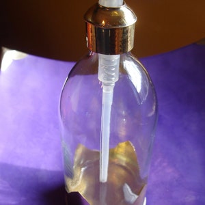 May include: A clear glass soap dispenser with a gold-coloured pump and base. The dispenser has a long white plastic tube and a decorative gold accent inside the bottle. The background is a gradient of purple and brown.