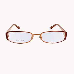 May include: Rectangular eyeglasses with a thin, reddish-brown metal frame. The lenses are clear, and the brand name "GUCCI" is printed on the left lens. The temples are a lighter brown colour.