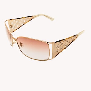 May include: Gold-framed sunglasses with gradient lenses. The temples feature a patterned design and are connected to the frame with a gold hinge. The lenses transition from a light pink to a darker shade.