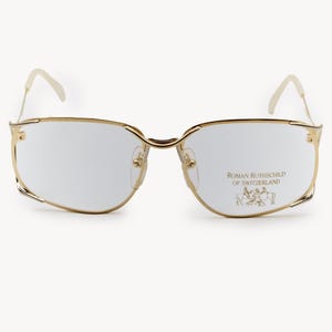 May include: Gold-rimmed spectacles with a rectangular lens shape. The glasses have a thin, delicate frame and temples. The text "ROMAN ROTHSCHILD OF SWITZERLAND" is printed on the right lens, along with a small emblem.