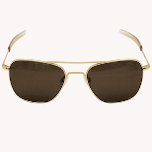 Puede incluir: Gafas de sol de aviador con montura dorada y lentes marrón oscuro. Las gafas tienen un doble puente y una forma clásica de lágrima. Las patillas son finas y doradas, con terminales transparentes. El diseño es elegante y atemporal.