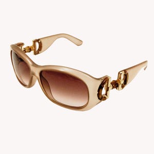 May include: Beige-toned sunglasses with brown gradient lenses. The frames have a rounded, oval shape and are accented with gold-colored metal details at the hinges. The temples are a matching beige color.