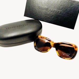 Può includere: Occhiali da sole tartarugati con lenti marroni, custodia nera e scatola nera. Gli occhiali da sole e la custodia sono impressi con il marchio "RALPH LAUREN". Gli occhiali hanno una montatura curva.