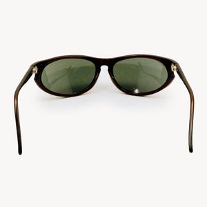 May include: Dark brown sunglasses with oval lenses and dark green tint. The sunglasses have a classic design with thin, curved arms and a subtle frame. The lenses are slightly reflective.
