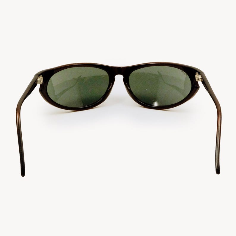 May include: Dark brown sunglasses with oval lenses and dark green tint. The sunglasses have a classic design with thin, curved arms and a subtle frame. The lenses are slightly reflective.
