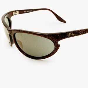May include: Brown sunglasses with a speckled pattern and gray lenses. The Ray-Ban logo is visible on the temple. The sunglasses have a curved design.
