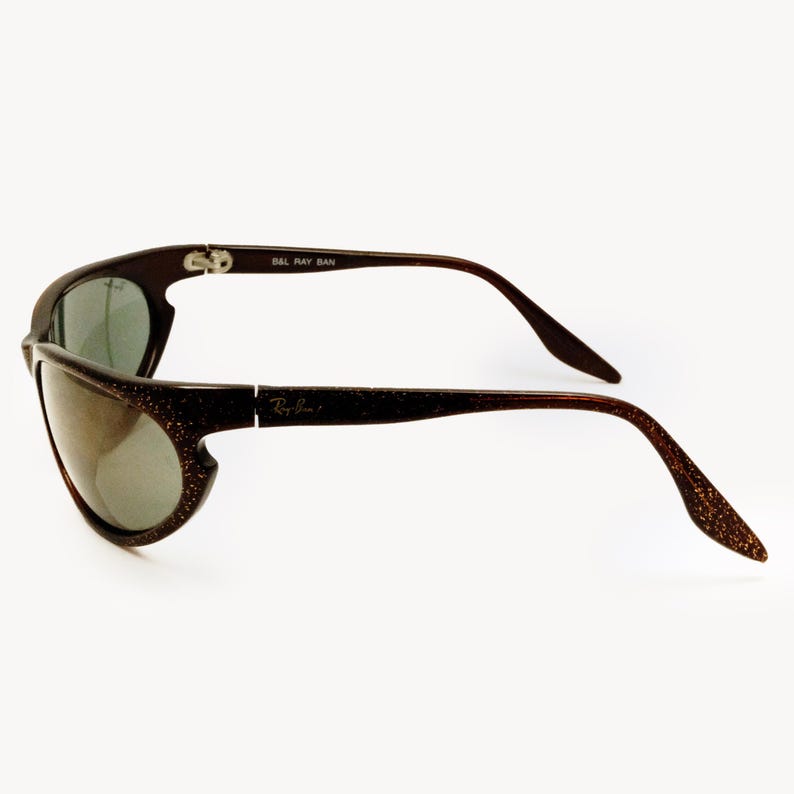 May include: Brown Ray-Ban sunglasses with a glittery finish. The sunglasses have a curved design with dark lenses and the Ray-Ban logo on the side. The text "B&L Ray Ban" is printed on the top of the frame.