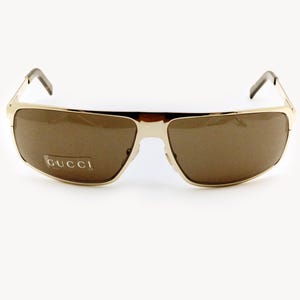 Puede incluir: Gafas de sol doradas con lentes marrones. Las gafas tienen forma rectangular y la marca "GUCCI" es visible en la lente izquierda. Las patillas son de color marrón oscuro.