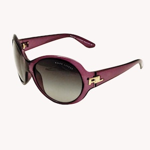 Puede incluir: Gafas de sol moradas con lentes degradadas. Las gafas tienen una montura gruesa con detalles dorados y las letras "RL". La marca "RALPH LAUREN" está impresa en la montura.