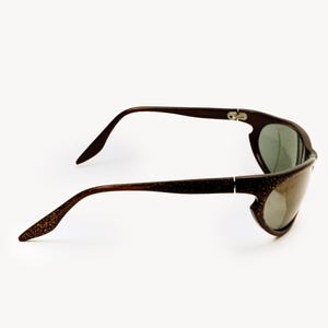 May include: Brown sunglasses with a glittery finish on the temples and a dark tint. The sunglasses have a unique, curved frame design and are set against a white background.