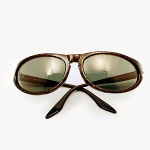 May include: Brown oval sunglasses with a speckled pattern on the frames and dark lenses. The arms are a solid brown color. The text "BAL MAY 5" is visible on the lens.