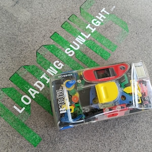 Op de afbeelding: Een kleurrijke speelgoedcamera met een rood, geel, blauw en groen ontwerp. De camera heeft een transparant kunststof behuizing en de tekst "XOBBOX" en "RICOH" zijn zichtbaar op de camera. De camera staat op een grijs oppervlak met groene tekst die "LOADING SUNLIGHT" luidt.