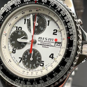 Peut inclure: Gros plan d'une montre chronographe Nismo argentée et noire. La montre présente un cadran blanc avec des sous-cadrans noirs, des aiguilles rouges et le logo Nismo. La lunette a des marquages noirs et blancs.