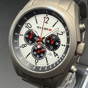 Pode incluir: Relógio de pulso prateado com mostrador branco, detalhes em preto e vermelho e o logotipo "nismo". O relógio possui vários mostradores secundários e uma luneta taquimétrica. Bracelete de metal e acabamento texturizado.