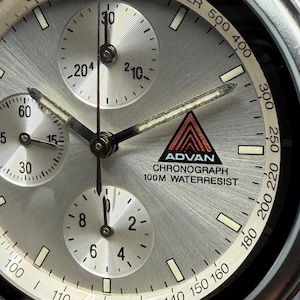 May include: Close-up of a silver ADVAN chronograph watch face. The watch features three sub-dials, a black and red logo, and the text "CHRONOGRAPH 100M WATERRESIST". The watch hands and markers are black.