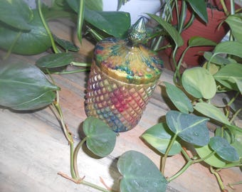 May include: A decorative, colorful glass jar with a diamond pattern and a pointed lid. The jar is gold, green, red, and blue. The jar is surrounded by green plants with heart-shaped leaves.