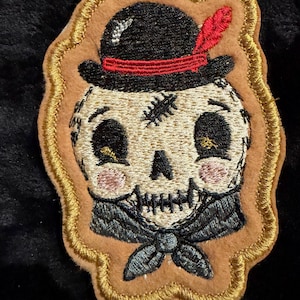 May include: An embroidered patch featuring a cream-colored skull wearing a black hat with a red band and feather. The skull has stitched details, pink cheeks, and a black neckerchief. The patch is outlined in gold thread on a tan felt base.