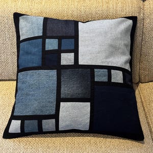 Peut inclure: Coussin carré décoratif avec un motif patchwork. Le coussin présente un motif géométrique de carrés en denim dans diverses nuances de bleu et de gris, soulignés de tissu noir. Le coussin mesure environ 45 cm de côté.