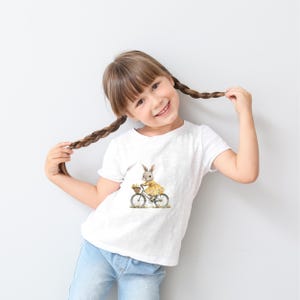May include: A white t-shirt with a graphic of a bunny riding a bicycle with a basket of flowers. The bunny is wearing a yellow dress.