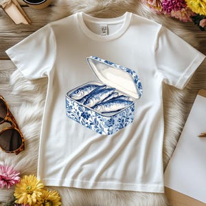 May include: White t-shirt with a blue and white illustration of a can of sardines. The can is open and the sardines are visible inside.