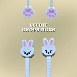 May include: A pair of white chopsticks featuring Leebit rabbit character heads with pink inner ears and black facial features, accompanied by two separate white paw-shaped attachments with pink pads. The text LEEBIT CHOPSTICKS is centered.