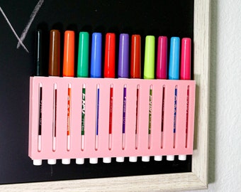 May include: A pink plastic holder with 12 colorful markers. The markers are arranged in a row and are labeled with different colors. The holder is attached to a black chalkboard.
