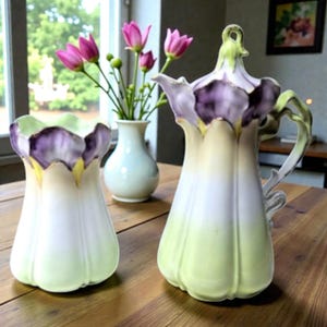 May include: A decorative ceramic pitcher and vase set. The pitcher has a handle and a flower-shaped lid. The set is painted in shades of green, white, and purple. The vase is a similar shape and color. A small white vase with pink flowers is in the background.