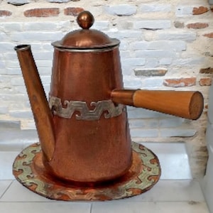 May include: A copper coffee pot with a matching circular tray. The pot features a long spout, a lid with a spherical knob, and a horizontal wooden handle. A silver-toned metal band with a repeating wave pattern encircles the pot and the tray.