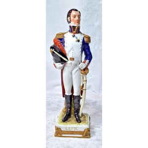 Puede incluir: Figura de porcelana de un oficial militar con uniforme blanco y azul, sosteniendo un sombrero y una espada. La figura tiene detalles dorados y está sobre una base con el nombre "LEPIC".