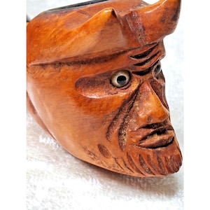 Puede incluir: Una pipa de madera tallada que presenta una cara detallada con cuernos, una nariz prominente y una barba tallada. La pipa está hecha de madera marrón claro con vetas visibles y tallas intrincadas, mostrando una estética rústica.