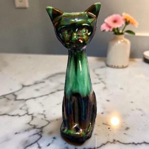 May include: A ceramic cat figurine with a glossy, iridescent finish. The cat is primarily green with darker hues of blue and brown. It has pointed ears and a long, slender neck. The figurine is a decorative item.