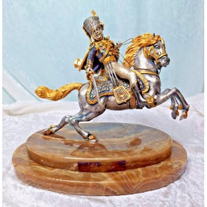 May include: A detailed figurine of a soldier on horseback. The sculpture features a silver horse with gold accents on its mane and hooves. The soldier wears a blue uniform with gold trim. The figurine is mounted on a two-tiered, brown marble base with the word "Votero".