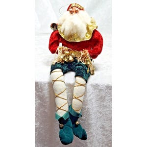 May include: A Santa Claus figurine wearing a red velvet top with a gold collar, dark green velvet shorts, and white leggings with gold crisscross ribbon detailing. The figure has a white beard, a red hat, and dark green pointed shoes with bells.