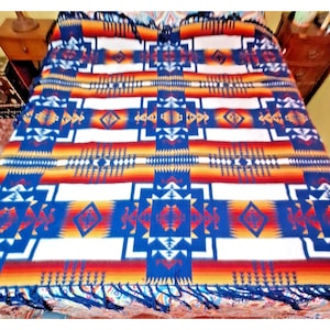 May include: A rectangular blanket featuring a repeating geometric pattern in shades of blue, white, orange, and red. The design includes diamond shapes, horizontal stripes, and stepped motifs. The edges are finished with dark blue fringe trim.