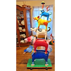 May include: A whimsical, colorful sculpture of dogs stacked on top of each other. The dogs are painted in bright colors: blue, red, white, yellow, and blue. The sculpture is on a green base with wheels. The top dog has a small umbrella.