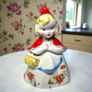 May include: A ceramic cookie jar in the shape of Little Red Riding Hood. The jar features a red hooded cape, a white dress with floral accents, and a yellow basket. The figure has blonde hair and is in a praying pose.