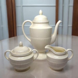 Puede incluir: Un juego de té de porcelana color crema, que incluye una cafetera alta con tapa, una azucarera con tapa y una crema. El juego presenta detalles acanalados verticales y un diseño similar a una cuerda alrededor de los bordes.
