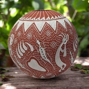 Puede incluir: Un jarrón de cerámica redondo, color terracota, con diseños blancos grabados. El jarrón presenta patrones geométricos, motivos florales y figuras estilizadas. El jarrón se exhibe sobre una superficie de madera con un fondo verde borroso.