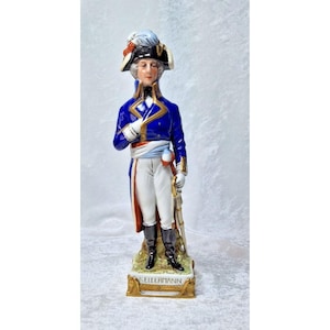 Op de afbeelding: Een porseleinen beeldje van een militair officier, gekleed in een blauwe jas met gouden biezen, een witte broek en een zwarte hoed met een veer. Het beeldje staat op een voetstuk met de naam "KELLERMANN" in gouden letters.