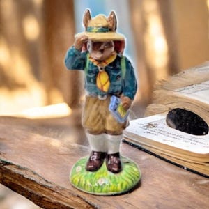 May include: A ceramic figurine of a rabbit dressed as a scout, wearing a tan hat, blue jacket, and khaki shorts. The rabbit is standing on a green base with grass, holding a map. The figurine is a charming collectible.