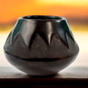 May include: A small, rounded black pottery vessel with a smooth, polished upper rim and a matte, textured lower section featuring a repeating triangular pattern around the circumference. The pot has a wide, open top and a tapered, bulbous body shape.