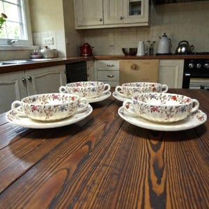 Op de afbeelding: Vier witte keramische soepkommen met bijpassende schotels. Elke kom en schotel is versierd met een bloemmotief in rood, blauw en roze. De kommen hebben twee handvatten en zijn gerangschikt op een donkere houten tafel.