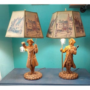 May include: Two vintage table lamps with shades depicting a town scene. The lamps feature a figure holding a lantern and a walking stick. The lampshades are square and the figures are brown. The lamps are on a blue surface.